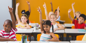 Students raise their hands and smile in classroom