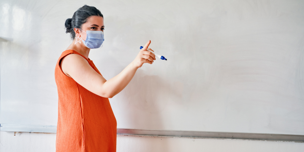 Pregnant teacher wearing face mask writes on whiteboard
